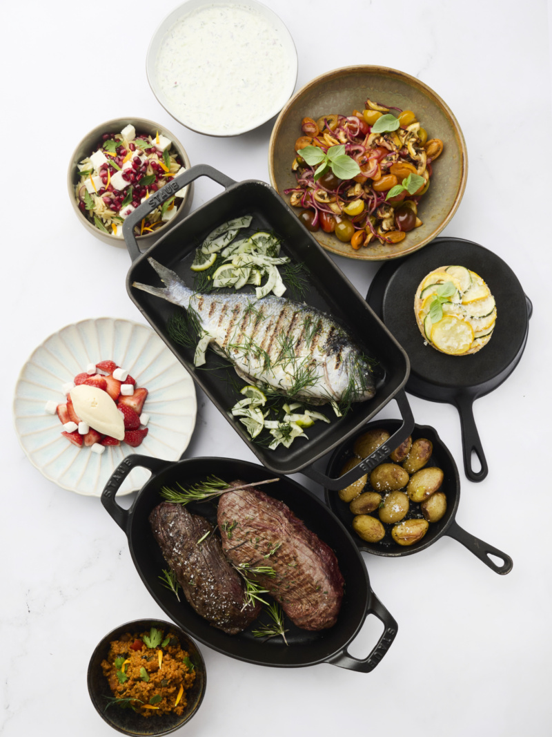 Luxe foodspread met gegrilde dorade, steaks, geroosterde aardappelen, couscous, tomatensalade, komkommersalade en aardbeiendessert, gepresenteerd in zwarte gietijzeren serveerpannen op een witte achtergrond.